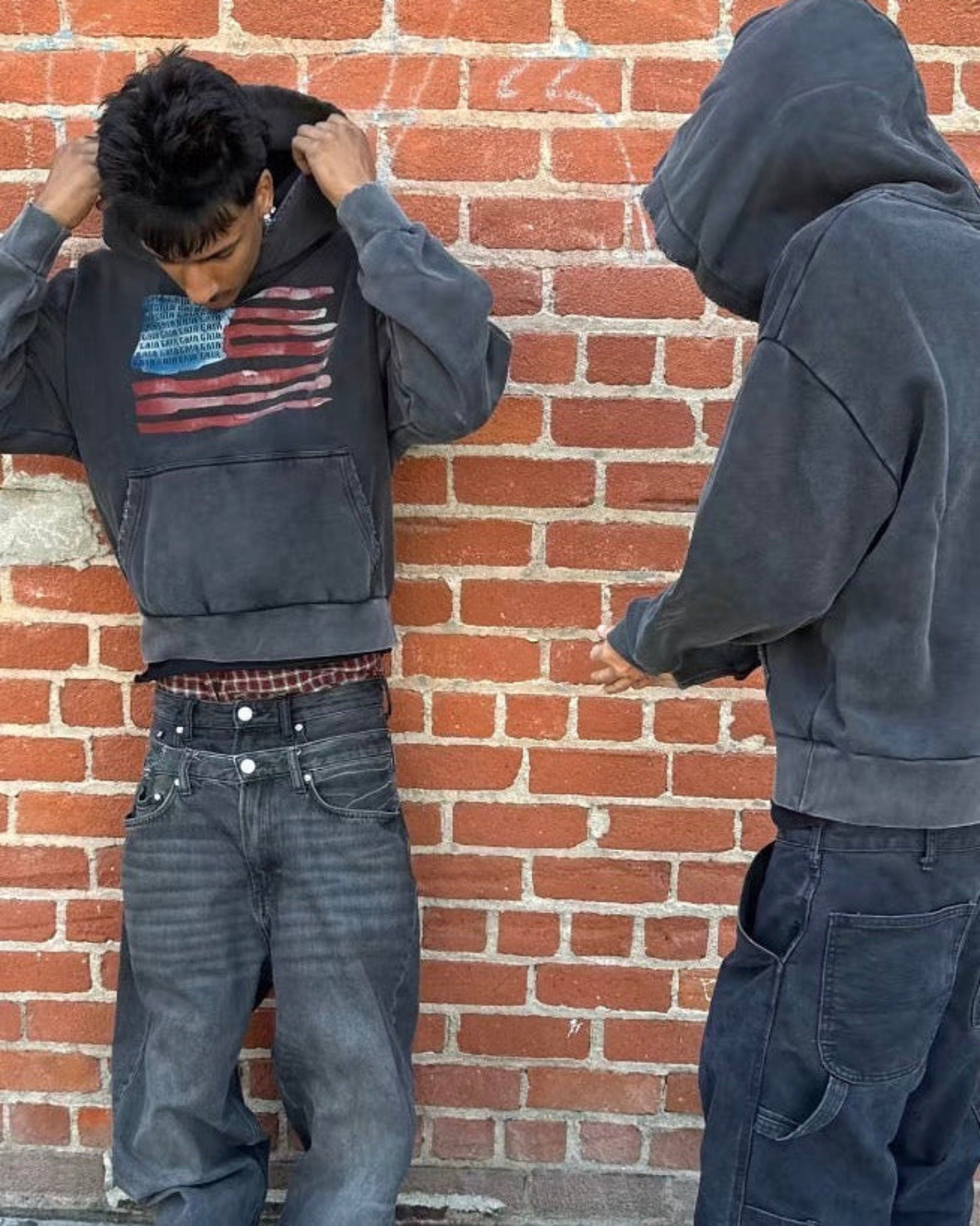 A person wearing a dark gray cropped hoodie with an American flag graphic, dark gray baggy jeans, and black shoes stands against a red brick wall. A second view on the right shows the back of the same person wearing the hoodie and dark pants.